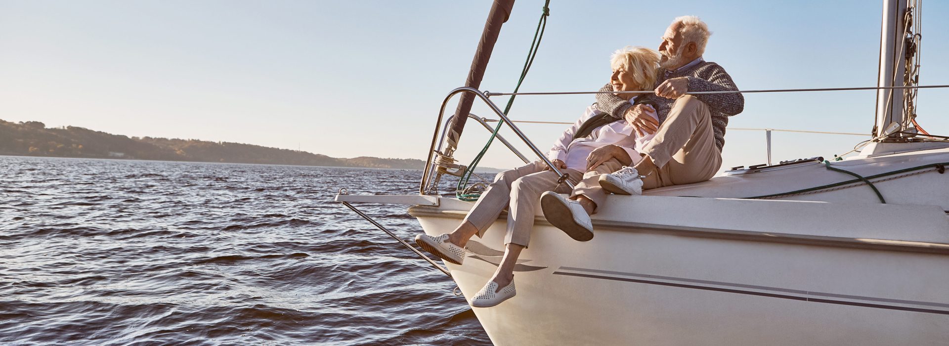 A happy senior couple sitting on the side of a sail boat on a calm blue sea. Man hugging his woman while enjoying view. Sailing, age, tourism, travel and people concept. Horizontal shot