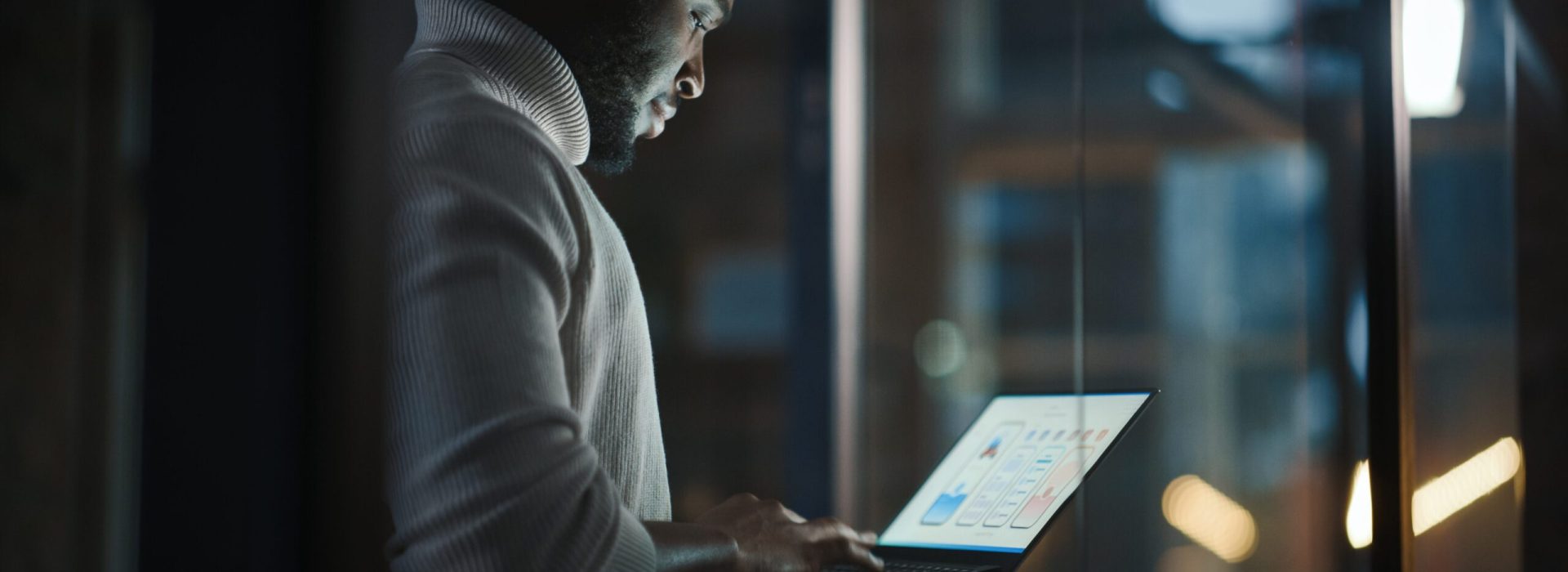 Handsome Black African American Male is Standing in Meeting Room Behind Glass Walls with Laptop Computer in an Creative Agency. Project Manager Wearing White Jumper and Working on App User Interface.